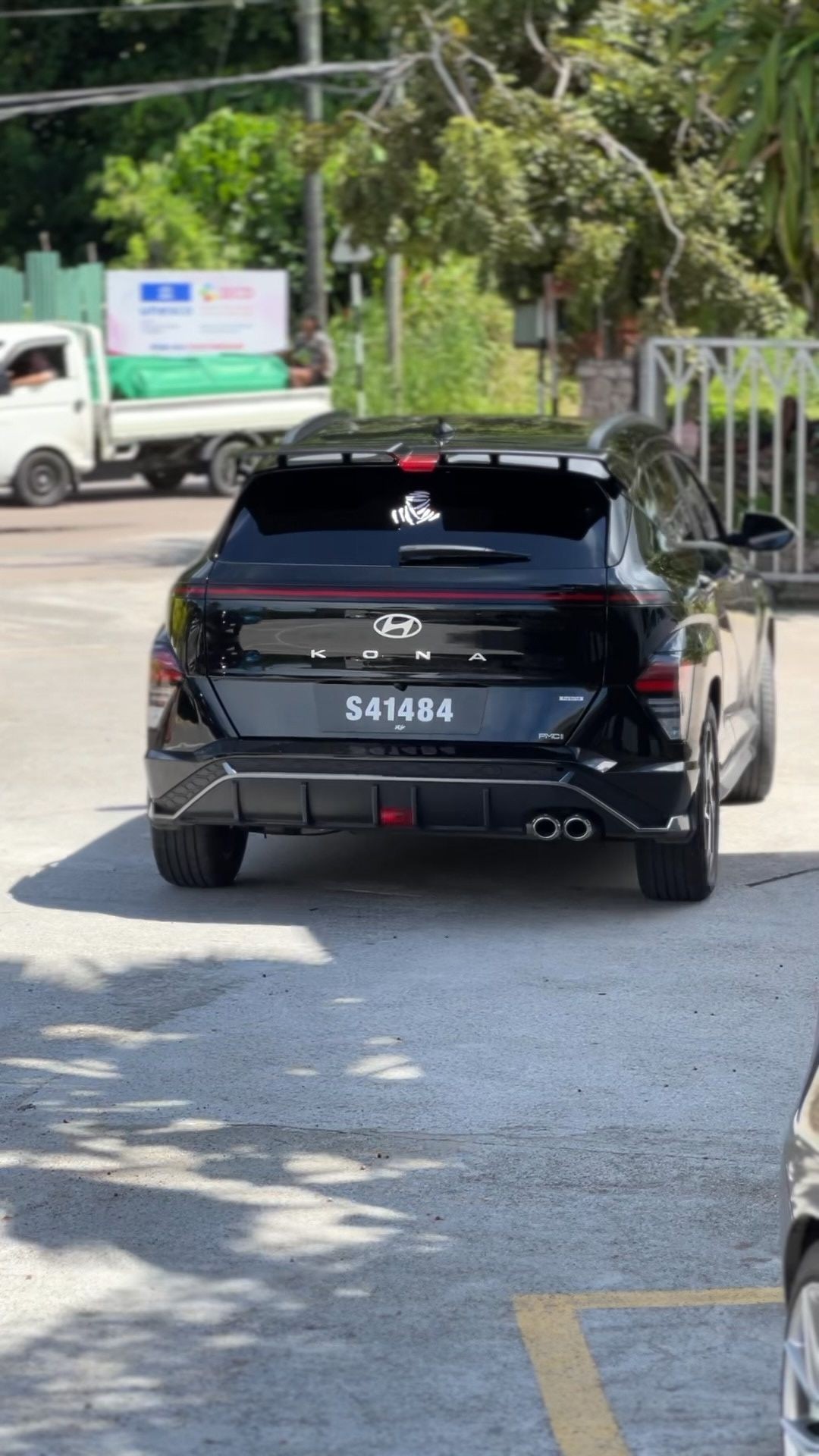 Black Hyundai Kona parked outdoors with trees in the background and a truck nearby.